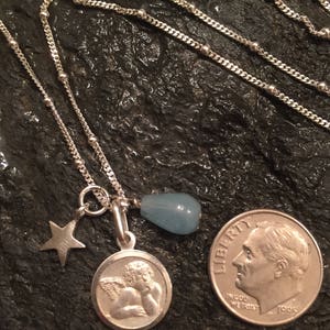 May include: A silver chain necklace with a charm featuring a cherub, a star, and a light blue teardrop-shaped stone. The necklace is layered on a dark grey textured surface.