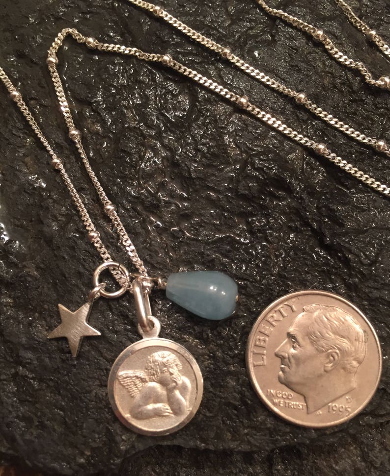 May include: A silver chain necklace with a charm featuring a cherub, a star, and a light blue teardrop-shaped stone. The necklace is layered on a dark grey textured surface.