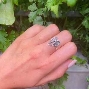 Peut inclure: Une bague en argent orn&eacute;e d'ailes d&eacute;taill&eacute;es avec un c&oelig;ur. La bague est pr&eacute;sent&eacute;e sur une main, avec un fond de verdure. Le design des ailes comprend des d&eacute;tails de plumes complexes, cr&eacute;ant un bijou d&eacute;licat et symbolique.