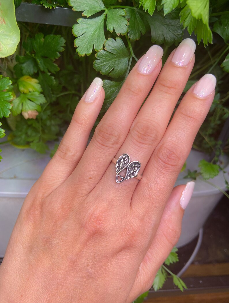 Peut inclure: Une bague en argent avec un motif en forme de c&oelig;ur et deux ailes. La bague est port&eacute;e sur une main avec du vernis &agrave; ongles rose clair. Le fond est une plante verte.