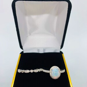 May include: A white opal engagement ring with a halo of smaller diamonds, and a matching wedding band with a delicate design of small diamonds. The rings are displayed in a black velvet ring box.
