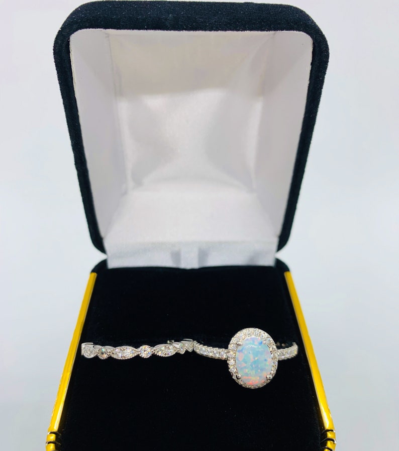 May include: A white opal engagement ring with a halo of smaller diamonds, and a matching wedding band with a delicate design of small diamonds. The rings are displayed in a black velvet ring box.