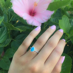 May include: A silver ring with a blue oval opal gemstone surrounded by a halo of smaller diamonds. The ring is on a hand with light pink nail polish.
