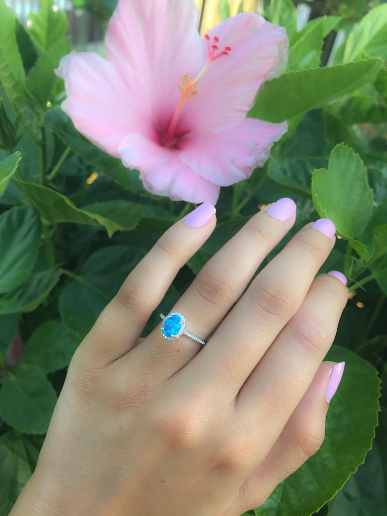 May include: A silver ring with a blue oval opal gemstone surrounded by a halo of smaller diamonds. The ring is on a hand with light pink nail polish.