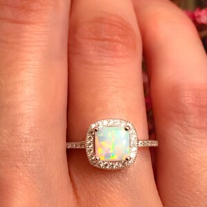 May include: A silver ring with a square-shaped opal gemstone surrounded by a halo of smaller diamonds.