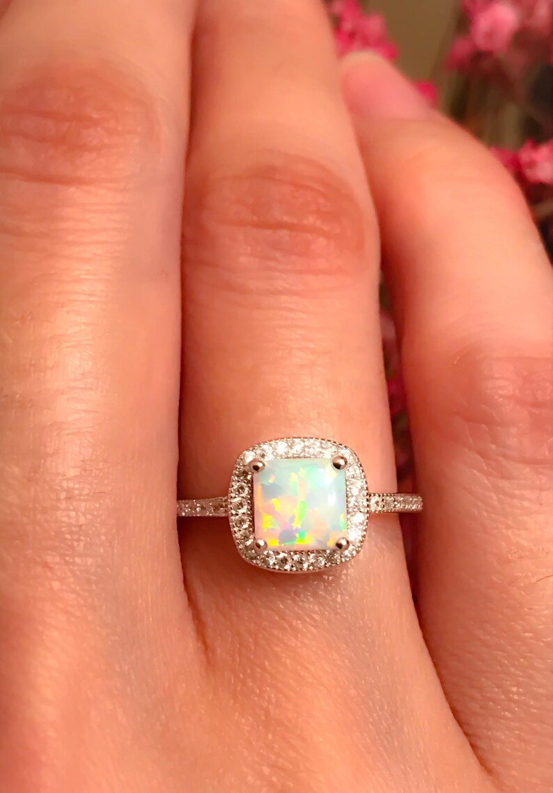 May include: A silver ring with a square-shaped opal gemstone surrounded by a halo of smaller diamonds.
