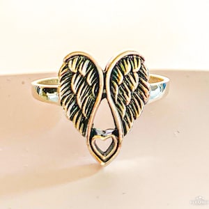 Peut inclure: Bague argent&eacute;e orn&eacute;e d'un motif en forme de c&oelig;ur avec des ailes d'ange. Les ailes pr&eacute;sentent des textures d&eacute;taill&eacute;es en forme de plumes, et une petite d&eacute;coupe en forme de c&oelig;ur est en bas. Sur fond neutre.