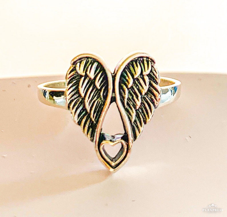 Peut inclure: Bague argent&eacute;e orn&eacute;e d'un motif en forme de c&oelig;ur avec des ailes d'ange. Les ailes pr&eacute;sentent des textures d&eacute;taill&eacute;es en forme de plumes, et une petite d&eacute;coupe en forme de c&oelig;ur est en bas. Sur fond neutre.