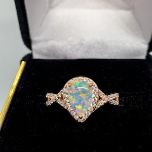 Op de afbeelding: Rosegouden ring met een peervormige opaal edelsteen. De opaal toont een levendig kleurenspel, omringd door een halo van kleine, heldere edelstenen. De ring heeft een delicaat, verweven bandontwerp, gepresenteerd in een zwarte sieradendoos.