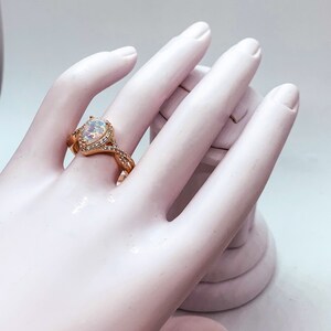 Op de afbeelding: Ros&eacute;gouden ring met een peervormige opaal middensteen, omringd door kleine, heldere edelstenen. De band heeft een gedraaid ontwerp met extra kleine edelstenen. De ring wordt getoond op een witte hand.