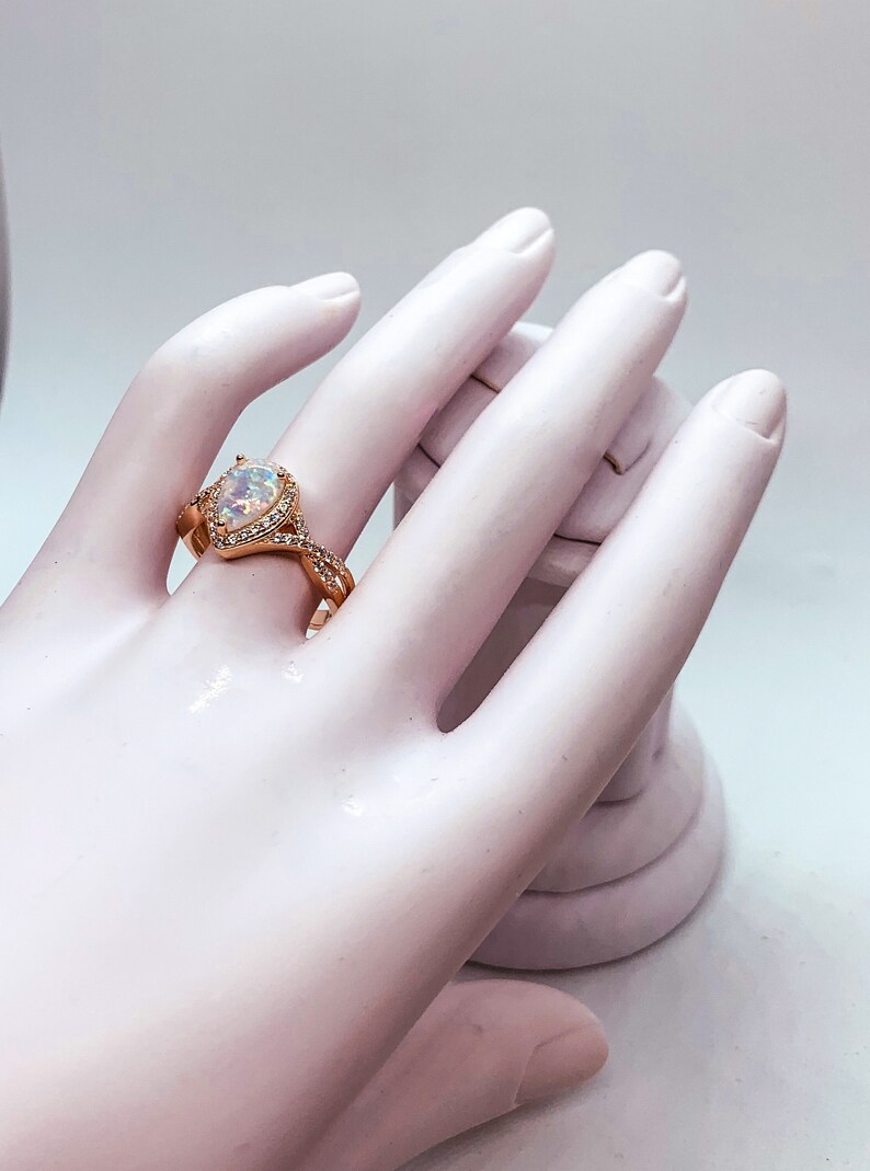 Op de afbeelding: Ros&eacute;gouden ring met een peervormige opaal middensteen, omringd door kleine, heldere edelstenen. De band heeft een gedraaid ontwerp met extra kleine edelstenen. De ring wordt getoond op een witte hand.