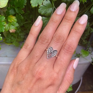 Peut inclure: Bague argent&eacute;e orn&eacute;e d'un c&oelig;ur et d'ailes d'ange. La bague est port&eacute;e au doigt, sur un fond de feuillage vert.
