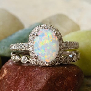 May include: A silver ring set with a large oval opal surrounded by a halo of smaller diamonds. The ring is on a brown rock.