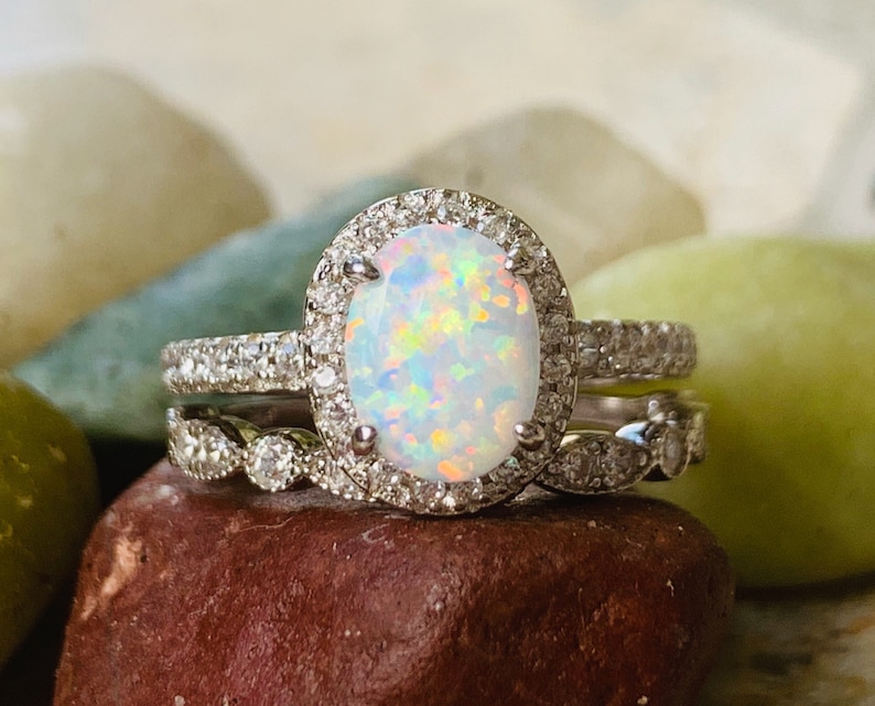 May include: A silver ring set with a large oval opal surrounded by a halo of smaller diamonds. The ring is on a brown rock.