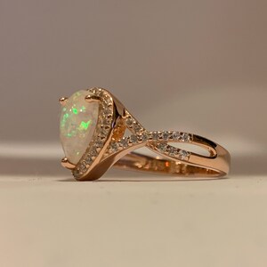 Op de afbeelding: Ros&eacute;gouden ring met een peervormige opaal edelsteen, omringd door een halo van kleine, heldere edelstenen. De band heeft een gedraaid ontwerp. De opaal heeft groene flitsen.