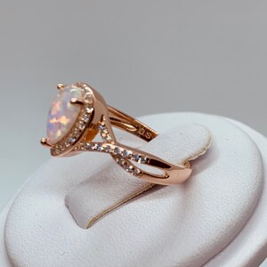 Op de afbeelding: Ros&eacute;gouden ring met een peervormige opaal edelsteen, omringd door kleine, heldere kristallen. De band heeft een gedraaid ontwerp, met extra kristallen. De ring wordt op een witte standaard getoond.