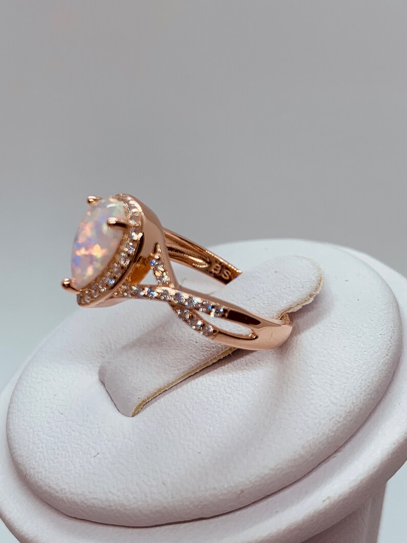 Op de afbeelding: Ros&eacute;gouden ring met een peervormige opaal edelsteen, omringd door kleine, heldere kristallen. De band heeft een gedraaid ontwerp, met extra kristallen. De ring wordt op een witte standaard getoond.