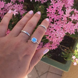 May include: A hand wearing three silver rings. The ring on the middle finger has a square blue stone surrounded by smaller stones. The other two rings are thin and delicate.