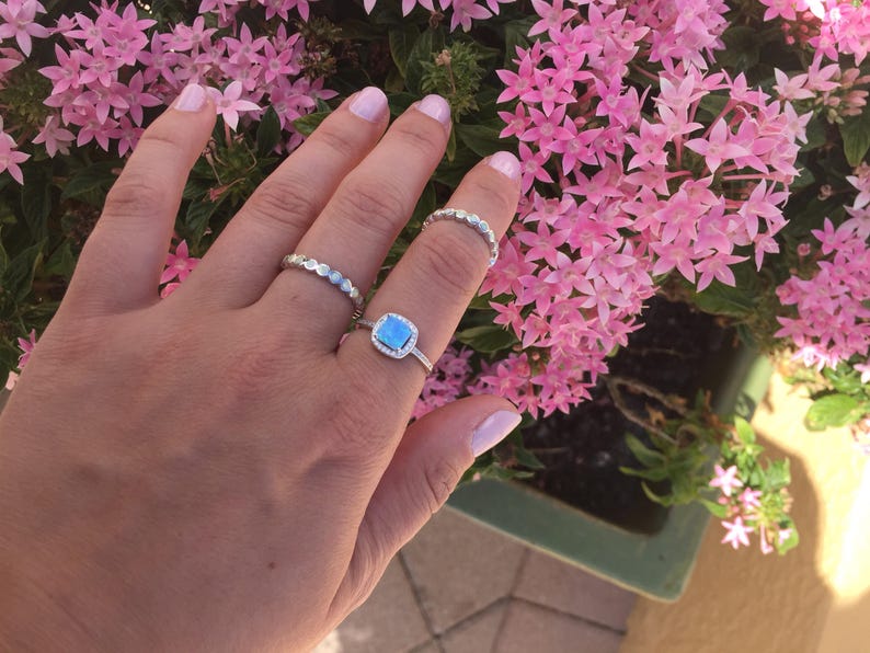 May include: A hand wearing three silver rings. The ring on the middle finger has a square blue stone surrounded by smaller stones. The other two rings are thin and delicate.