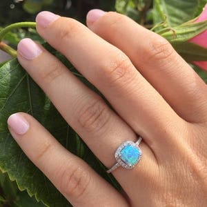 May include: A silver ring with a square-shaped blue opal gemstone surrounded by a halo of smaller clear gemstones. The ring is on a finger.