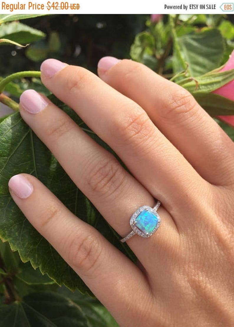 May include: A silver ring with a square-shaped blue opal gemstone surrounded by a halo of smaller clear gemstones. The ring is on a finger.