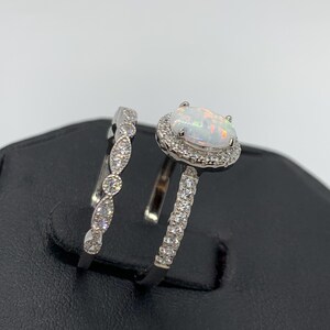 May include: A silver ring set with a white opal center stone surrounded by a halo of smaller stones. The matching band features a delicate, curved design with small stones.