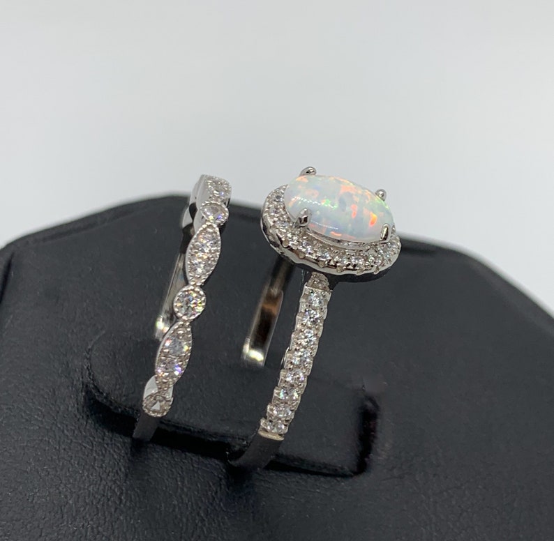 May include: A silver ring set with a white opal center stone surrounded by a halo of smaller stones. The matching band features a delicate, curved design with small stones.