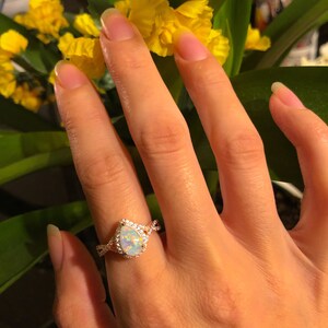 Op de afbeelding: Een ros&eacute;gouden ring met een peervormige opaal als middensteen, omringd door een halo van kleine, heldere edelstenen. De band heeft een gedraaid ontwerp. De ring zit aan een hand, met gele bloemen op de achtergrond.
