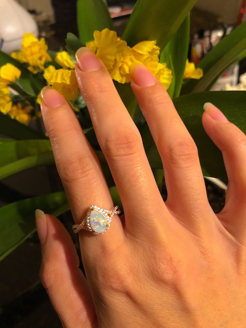 Op de afbeelding: Een ros&eacute;gouden ring met een peervormige opaal als middensteen, omringd door een halo van kleine, heldere edelstenen. De band heeft een gedraaid ontwerp. De ring zit aan een hand, met gele bloemen op de achtergrond.