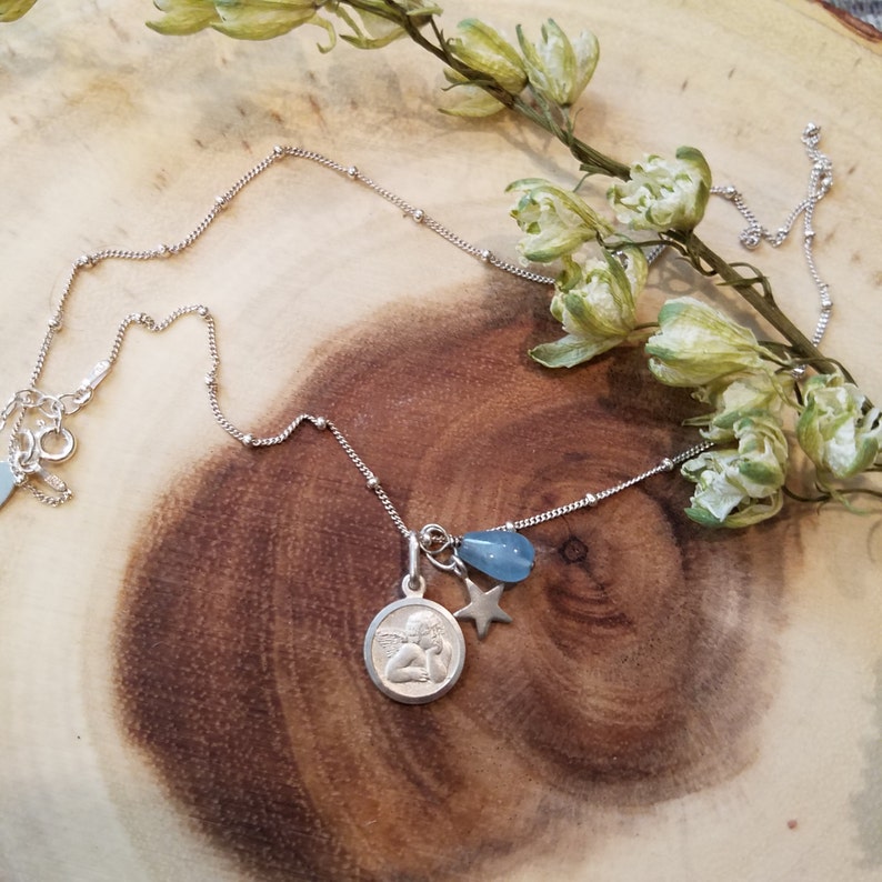 May include: A silver necklace with a delicate chain and a charm featuring an angel with a blue teardrop-shaped stone and a silver star.