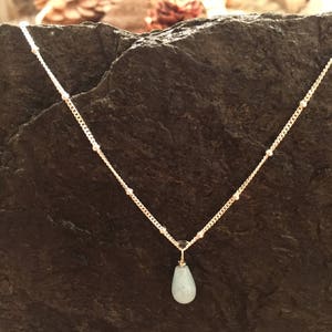 May include: A delicate gold chain necklace with a light blue teardrop pendant. The necklace is on a dark grey rock background.