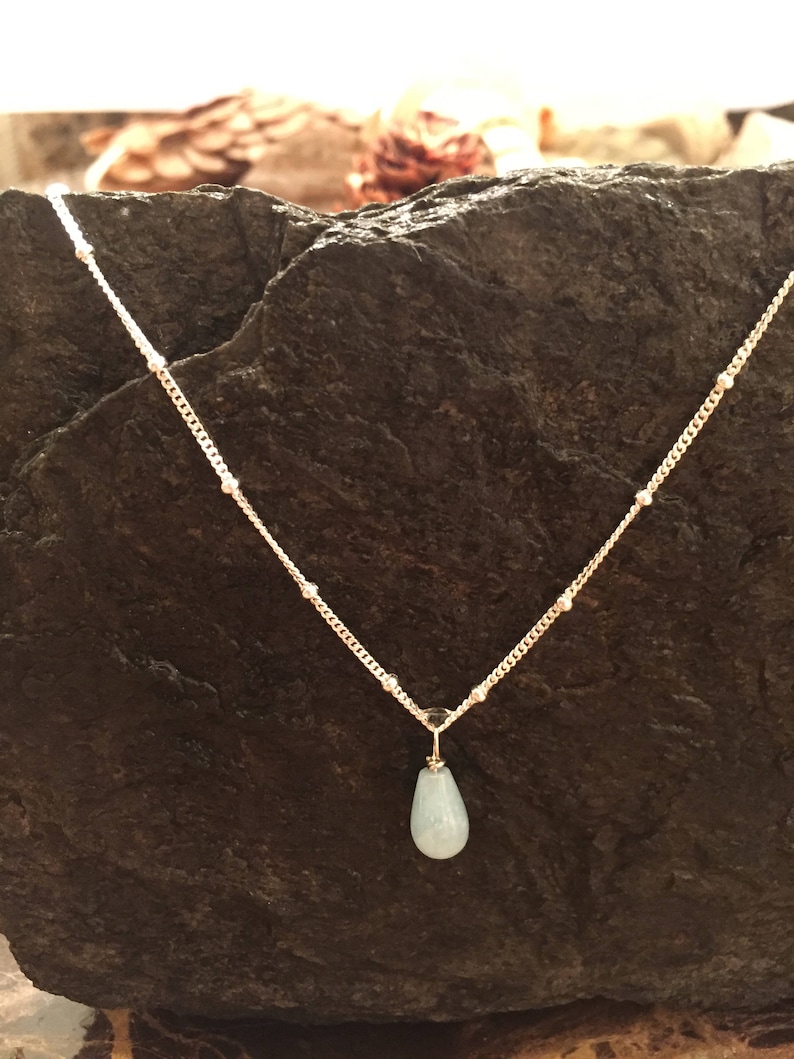 May include: A delicate gold chain necklace with a light blue teardrop pendant. The necklace is on a dark grey rock background.