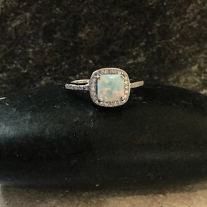 May include: A silver ring with a square-shaped opal gemstone surrounded by a halo of smaller diamonds.