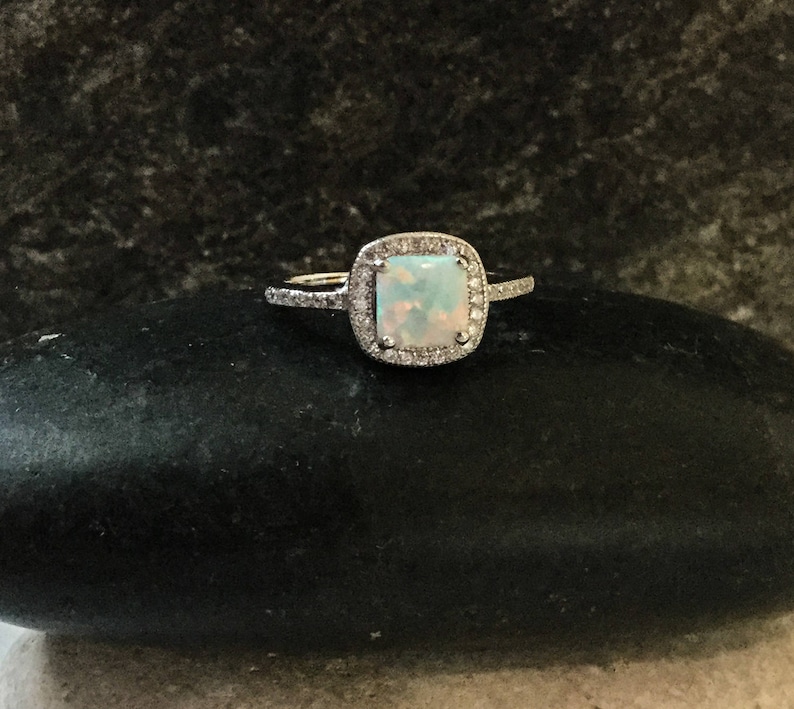 May include: A silver ring with a square-shaped opal gemstone surrounded by a halo of smaller diamonds.