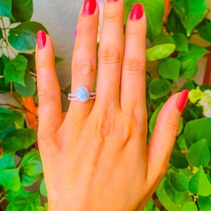 May include: A silver ring with a large oval opal gemstone surrounded by smaller clear gemstones. The ring is on a person's finger with red nail polish.