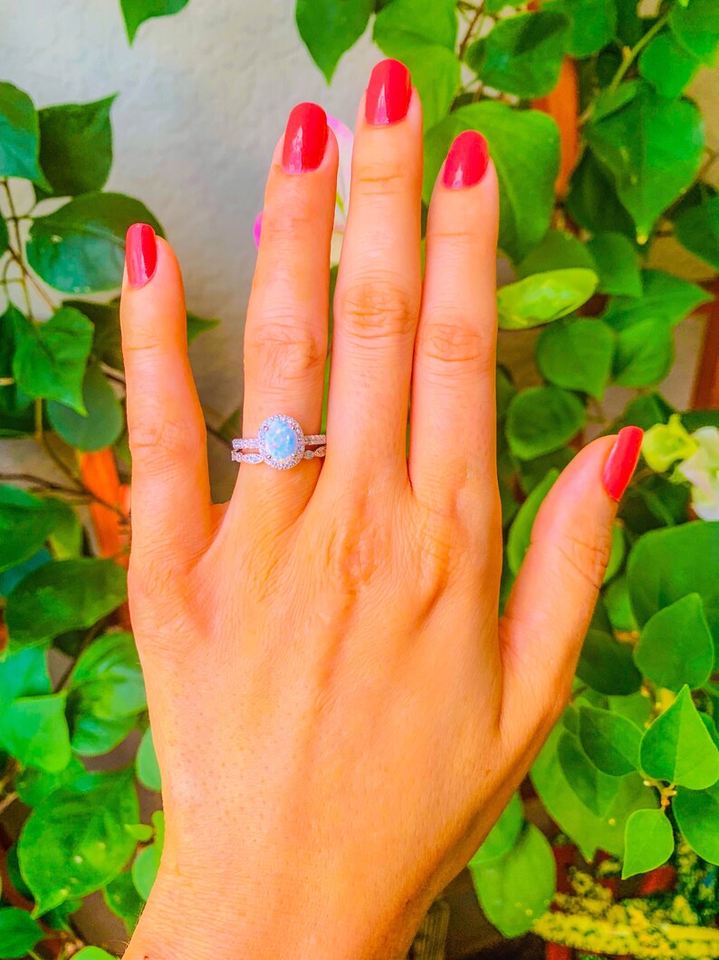 May include: A silver ring with a large oval opal gemstone surrounded by smaller clear gemstones. The ring is on a person's finger with red nail polish.