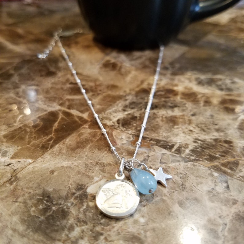 May include: A silver chain necklace with a round pendant featuring an angel design. The pendant is attached to a small blue teardrop-shaped gemstone and a silver star charm.