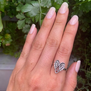 Peut inclure: Bague en argent repr&eacute;sentant un c&oelig;ur avec des ailes d'ange. La bague est port&eacute;e sur une main avec du vernis &agrave; ongles rose clair. Le fond montre des feuilles vertes.