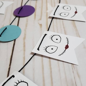 Pu&ograve; includere: Una ghirlanda di corda nera con bandierine di carta bianca che presentano illustrazioni in bianco e nero del viso di un personaggio con dettagli cuciti. La ghirlanda include anche cerchi viola e blu.