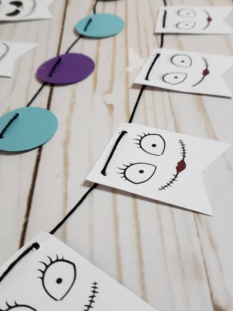 Pu&ograve; includere: Una ghirlanda di corda nera con bandierine di carta bianca che presentano illustrazioni in bianco e nero del viso di un personaggio con dettagli cuciti. La ghirlanda include anche cerchi viola e blu.