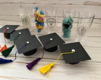 May include: A selection of graduation party favours. Features black mortarboard hats with coloured tassels, clear plastic cups with graduation-themed text and images, and sweets.