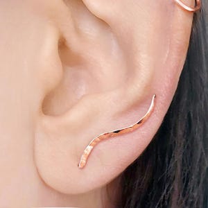 Puede incluir: Primer plano de una oreja adornada con dos joyas. Un pequeño trepador de oreja de oro rosa martillado se curva a lo largo del borde de la oreja. También es visible un pendiente de aro de oro rosa.