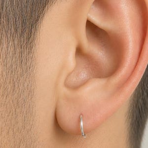 Può includere: Primo piano di un orecchio umano con un piccolo orecchino a cerchio argentato. L'orecchino è una sottile fascia semplice. La pelle intorno all'orecchio è liscia e chiara. Lo sfondo è sfocato, focalizzando l'attenzione sull'orecchino.