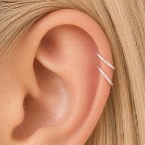 May include: Close-up of an ear with two small, silver hoop earrings. The earrings are simple and elegant, adding a subtle touch of style. The ear is set against a backdrop of blonde hair.