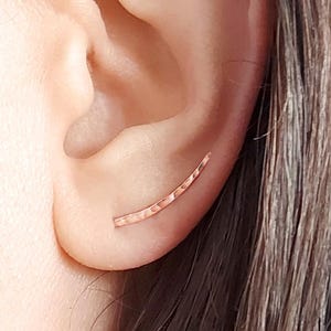 Puede incluir: Primer plano de una oreja con dos joyas. Un pendiente pequeño y curvo de metal martillado está en la parte inferior de la oreja. Un pequeño pendiente de aro dorado está en la parte superior de la oreja.