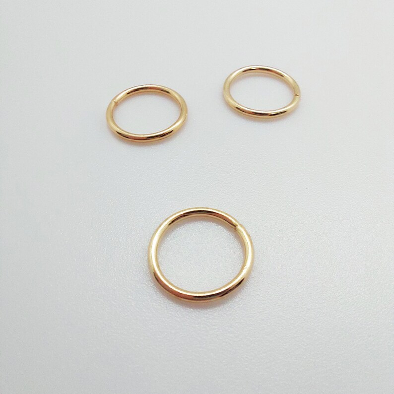 Puede incluir: Tres pendientes de aro circulares de color dorado. Los pendientes est&aacute;n hechos de un metal liso y brillante y son de diferentes tama&ntilde;os. Los aros se muestran sobre una superficie blanca.