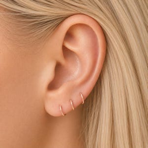 Puede incluir: Primer plano de una oreja con tres pendientes de aro peque&ntilde;os y delicados en oro rosa. Los pendientes est&aacute;n espaciados uniformemente y crean un look minimalista y elegante. La oreja est&aacute; sobre un fondo de pelo rubio.