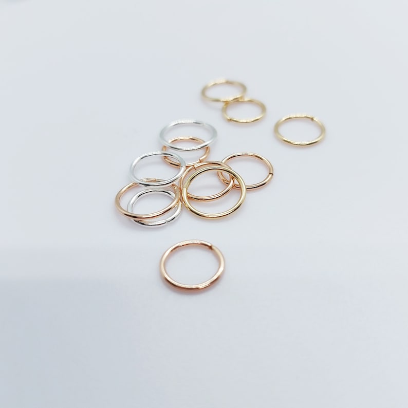 Puede incluir: Una colecci&oacute;n de peque&ntilde;os y finos pendientes de aro en varios acabados met&aacute;licos, incluyendo oro, plata y oro rosa. Los pendientes est&aacute;n dispersos sobre una superficie blanca, mostrando su dise&ntilde;o circular y sencillo. Adecuados para el uso diario.