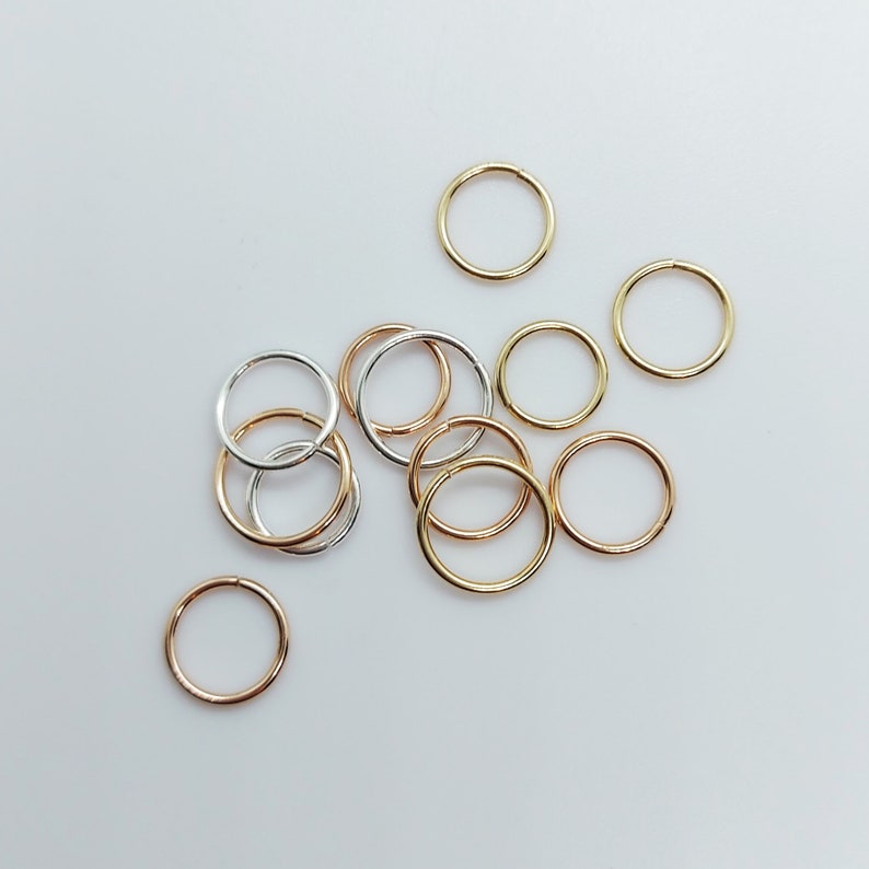 Puede incluir: Una colecci&oacute;n de peque&ntilde;os pendientes circulares en varios acabados met&aacute;licos, incluyendo oro, plata y oro rosa. Los pendientes est&aacute;n dispersos sobre una superficie blanca, mostrando su dise&ntilde;o simple y minimalista. La imagen destaca las diferentes opciones de color disponibles.
