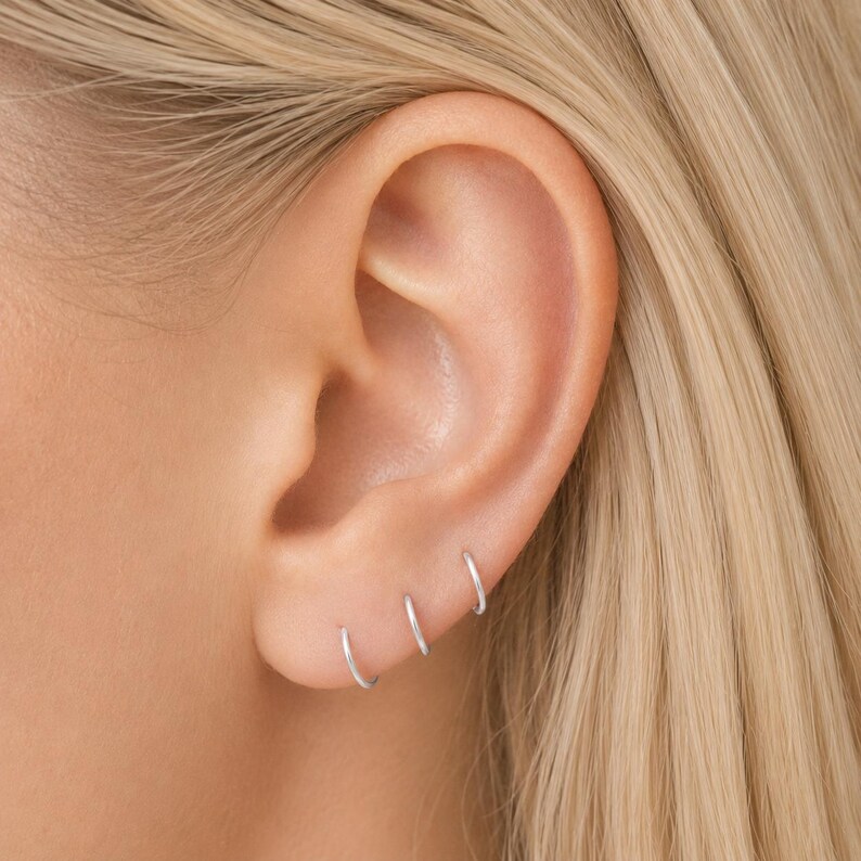 Puede incluir: Primer plano de una oreja con tres peque&ntilde;os pendientes de aro plateados. Los pendientes son sencillos y minimalistas. La oreja est&aacute; contra el pelo rubio.